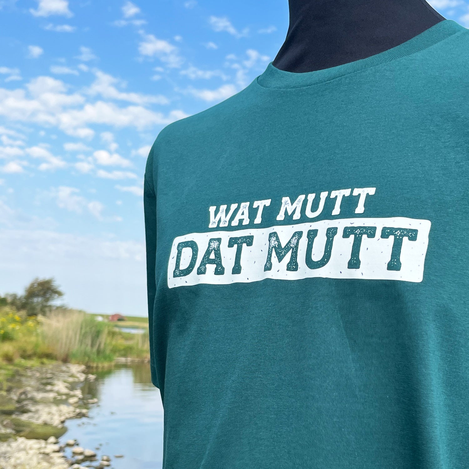 Grünes Unisex T-Shirt mit Rundhalsausschnitt. Auf der Brust steht in weiß der Spruch Wat mutt dat mutt. Die Farbe vom T-Shirt heißt Glazed Green. Im Hintergrund ist eine typische Norddeutsche Landschaft zu sehen.