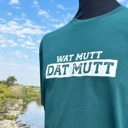 Grünes Unisex T-Shirt mit Rundhalsausschnitt. Auf der Brust steht in weiß der Spruch Wat mutt dat mutt. Die Farbe vom T-Shirt heißt Glazed Green. Im Hintergrund ist eine typische Norddeutsche Landschaft zu sehen.
